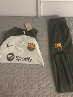 Barcelona träningsset Nike grön/vit - Snyggt träningsset från Nike och FC Barcelona med långärmad tröja i vitt och grönt samt matchande gröna byxor. Tröjan har halv dragkedja, klubbmärke och Spotify-logga på bröstet. Byxorna har resår i midjan och klubbmärke på benet. Perfekt för fotbollsträning.
