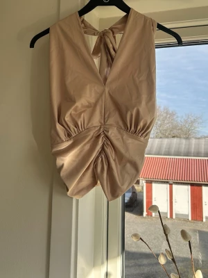 Champagnefärgad topp med knyte - Snygg champagnefärgad topp med djup v-ringning och knyt i nacken. Toppen har en draperad effekt framtill och är ärmlös, vilket ger en elegant och trendig look. Perfekt för dig som vill sticka ut lite extra.💛(liten fläck på framsidan längre ner)