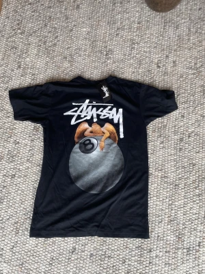 Svart Stüssy t-shirt med tryck - Svart t-shirt från Stüssy med stort grafiskt tryck på ryggen som visar en figur på en biljardboll och vit Stüssy-logga. Framtill finns en mindre logga på bröstet. Klassisk passform och rund hals. Perfekt för dig som gillar streetwear.