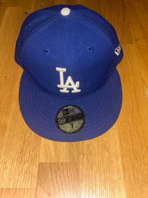 Blå LA Dodgers keps från New Era - Snygg blå keps från New Era med LA Dodgers-logga broderad framtill i vitt. Klassisk 59FIFTY-modell med platt skärm och broderade ventilationshål. Materialet är tåligt och kepsen har en ikonisk streetwear-look. Perfekt för dig som gillar sportig stil.