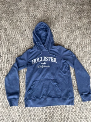 Blå hoodie från Hollister XS - Blå hoodie från Hollister med vit logga och texten 'California' på bröstet. Klassisk känguruficka framtill och justerbar huva. Mjuk insida i fleece och ribbade muddar vid ärmar och nederkant. Perfekt för en avslappnad och cool stil.
