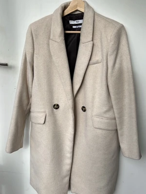 Beige kappa  - Stilren, ordentlig beige kappa i polyester & ull med två tunna axelvaddar, dubbelknäppning och fickor. Den går att sprätta upp baktill men är ej gjort så den är stängd hela vägen ner. På mig slutar den precis ovanför knäna, jag är 172 cm. 