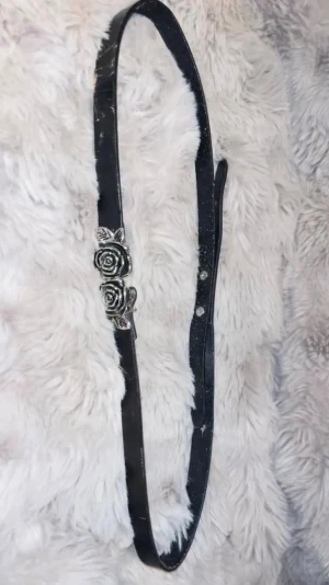 Svart bälte med rosdetalj i silver - Snyggt svart bälte med en unik spänne i form av två silverfärgade rosor. Bältet är smalt och passar perfekt till jeans eller klänning för att ge en cool touch till din outfit. Materialet är skinn och spännet ger en extra detalj som sticker ut.