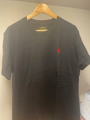 Svart t-shirt från Polo Ralph Lauren - Klassisk svart t-shirt från Polo Ralph Lauren med rund halsringning och det ikoniska röda broderade logot på bröstet. Utan defekter perfekt till sommardagarna 