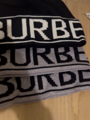 Burberry mössa i svart och grått - Säljer en snygg mössakfrån Burberry med stor logotyp i svart och vitt samt grått och svart. Halsduken är stickad och har en modern, grafisk design som sticker ut. Perfekt accessoar för att lyfta din outfit.