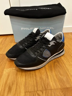 Philippe Model svarta sneakers med blå detalj - Snygga svarta sneakers från Philippe Model med blå och grå detaljer. Skorna är använda sparsamt då de ej varit helt rätt i. Finns en defekt på höger skon. Kontakta mig för mer info!                                                             Skorna levereras med originalkartong och dustbag.