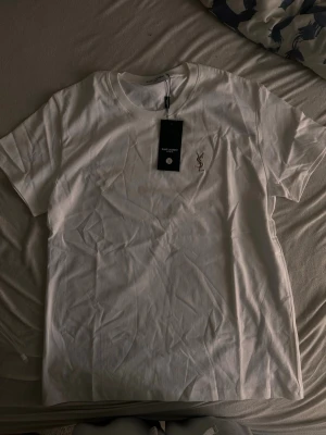 Vit t-shirt från Saint Laurent - Säljer en klassisk vit t-shirt från Saint Laurent med diskret YSL-logga broderad på bröstet. T-shirten har rund halsringning och korta ärmar. Tillverkad i mjuk bomull för en skön känsla och clean look.