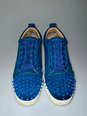 Blåa sneakers med nitar från Christian Louboutin  - Unika blåa sneakers från Christian Louboutin i mocka med matchande blå nitar på tån och blå skosnören. Skorna har vit sula och låg profil. Perfekta för dig som vill sticka ut med en edgy och cool stil.
