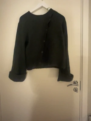 Svart kofta från Gina Tricot - Mysig svart kofta från Gina Tricot med knäppning framtill och rund halsringning. Koftan har långa ärmar med uppvikta muddar och en rak, något kortare passform. Perfekt för lager på lager och enkel att styla till olika outfits.