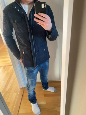 Marin-Blå quiltad jacka med dragkedja - Snygg quiltad jacka med dragkedja och knappar i guld. Jackan har flera fickor framtill och en hög krage. Perfekt för höst och vår, med en stilren och modern look.