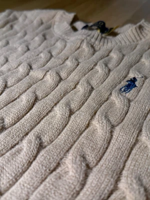 Beige kabelstickad tröja Polo Ralph Lauren - Kabelstickad tröja i beige från Polo Ralph Lauren med klassisk blå logga broderad på bröstet. Tröjan har rund halsringning och ribbade muddar. Perfekt för dig som gillar tidlös och stilren design.