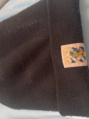Mörkblå ribbstickad mössa med patch - Säljer en mörkblå ribbstickad mössa med uppvikt kant och en liten patch med blått och gult mönster framtill. Klassisk och enkel design som passar till det mesta. Perfekt för kalla dagar och enkel att matcha med olika outfits. Forza Blåvitt 