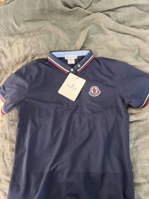 Marinblå pikéskjorta från Moncler - Snygg marinblå pikéskjorta från Moncler med klassisk krage och knappar framtill. Kragen och ärmsluten har röd, vit och blå randig detalj. Ikonisk Moncler-logga på bröstet. Perfekt för en stilren och sportig look. Helt ny!