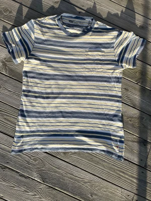  Vintage Randig Levis t-shirt 2000s  - Snygg randig t-shirt från Blend i blått, vitt och gult. T-shirten har korta ärmar, rund halsringning och en liten bröstficka. Perfekt för dig som gillar en avslappnad och fräsch stil. Tillverkad i mjuk bomull för skön känsla.