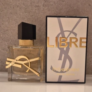 Yves Saint Laurent Libre Eau de Parfum - Säljer en elegant parfym från Yves Saint Laurent, Libre Eau de Parfum. Flaskan är transparent med guldiga detaljer och har en svart, kantig kork. Designen är lyxig med YSL-loggan i guld på flaskan och en strukturerad guldfärgad ring runt halsen.