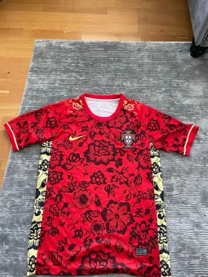 Portugal Nike röd  - Snygg Portugal matchtröja från Nike i rött med svart blommönster och gula detaljer. Tröjan har rund hals, korta ärmar och sidopaneler med blommigt mönster i gult och svart. Perfekt för dig som vill sticka ut på planen eller på läktaren.