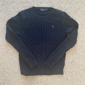 Mörkblå kabelstickad tröja Polo Ralph Lauren - Kabelstickad mörkblå tröja från Polo Ralph Lauren med rund halsringning och röd broderad logga på bröstet. Tröjan har långa ärmar och ribbade muddar vid ärmslut och nederkant. Perfekt för en klassisk och stilren look.