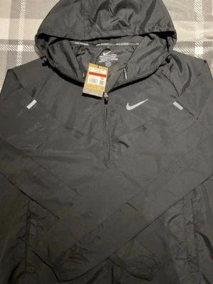 Svart Nike vindjacka med huva - Snygg svart vindjacka från Nike med huva och halv dragkedja framtill. Jackan har reflekterande detaljer på bröstet och ärmarna samt en klassisk Nike-logga. Tillverkad i lätt och slitstarkt syntetmaterial, perfekt för blåsiga dagar.