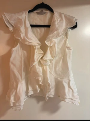 Vit volangblus med kort ärm - Snygg vit blus med volangdetaljer framtill och längs nederkanten. Blusen har korta ärmar och en luftig, feminin passform. Perfekt för dig som gillar romantiska detaljer och en lätt, somrig känsla. Materialet känns mjukt och svalt.