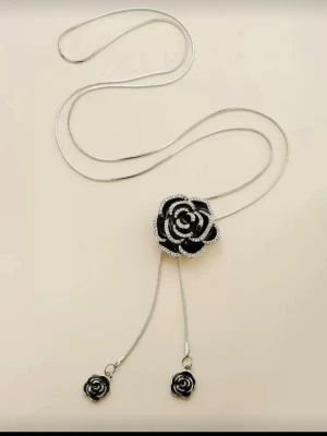 Glittrande Silverfärgat halsband med svart ros - Unikt halsband i silverfärg med en stor svart ros dekorerad med gnistrande stenar. Kedjan har två mindre rosor som detaljer i ändarna. Perfekt för dig som vill sticka ut med en cool och elegant accessoar. Materialet ser ut att vara metall och emalj.