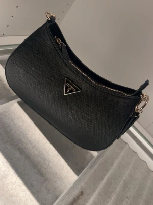 Svart axelväska från Guess - Snygg svart axelväska från Guess med guldfärgade detaljer och klassisk trekantslogga framtill. Väskan har justerbar axelrem och dragkedja både bak. 