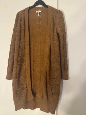 Brun lång kofta från Object - Mysig lång kofta i brun färg från Object. Koftan är stickad med ett snyggt hålmönster och har ribbade muddar vid ärmslut och nederkant. Perfekt att slänga över en t-shirt eller topp för extra värme och stil.