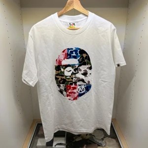 Bape T-shirt ~ Multicolor - White T-shirt in a classic fit 🔷 🔷 Iconic graphic print from A Bathing Ape featuring the signature Ape Head in a multicolor camouflage design 🔷 Bold statement front combining several camo patterns for a unique look 🔷 Clean white base that makes the graphic stand out 🔷 Soft cotton fabric for everyday comfort 🔷 Ribbed crewneck collar for a classic and comfortable fit 🔷 Easy to style – pairs well with both casual and streetwear outfits 🔷 Recognizable streetwear piece with one of BAPE’s most iconic 