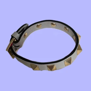 Valentino armband med nitar i guld - Snyggt armband från Valentino i ljusgrått skinn. Justerbart spänne i guld och svart kant. Använts men är fortfarande i bra skick!! 
