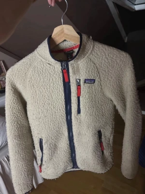 Beige fleecejacka från Patagonia - Säljer en beige, fluffig fleecejacka från Patagonia med marinblå detaljer och röda dragkedjor. Jackan har hel dragkedja framtill, tre fickor med dragkedja och en liten logga på bröstet. Perfekt för kalla dagar och riktigt mysig att ha på sig. Har ett liten hål vid slutet av ärmen men kan säkert lätt sys, förutom det lite nopprig typ. Annars är skicket bra. 
