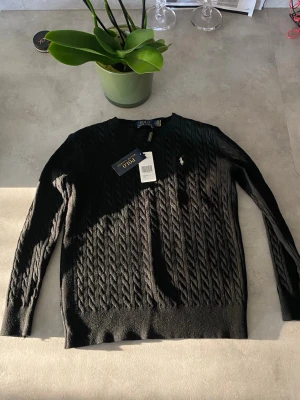 Svart kabelstickad tröja Polo Ralph Lauren - Snygg svart kabelstickad tröja från Polo Ralph Lauren med rund halsringning och den klassiska broderade loggan på bröstet. Tröjan har långa ärmar och är tillverkad i mjukt material som passar perfekt till höst och vinter.