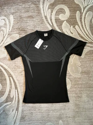 Svart träningströja från Gymshark - Snygg svart träningströja från Gymshark med gråa detaljer och meshmönster upptill. I storlek M, Tight passform och korta ärmar, perfekt för gymmet eller löpning. Tillverkad i ett stretchigt funktionsmaterial som andas och transporterar bort fukt.