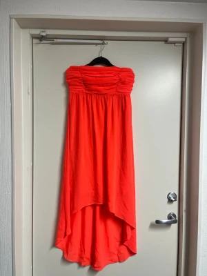 Axelbandslös orange långklänning - Snygg axelbandslös långklänning i stark orange färg med rynkad överdel och asymmetrisk nederkant. Perfekt för sommarens alla tillfällen. Klänningen har en flowy passform och är tillverkad i ett lätt material.