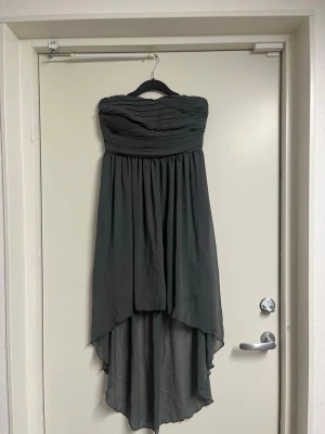 Mörkgrå axelbandslös klänning - Säljer en elegant mörkgrå axelbandslös klänning med draperad överdel och asymmetrisk nederkant. Klänningen har en flowy passform och är perfekt för dig som vill ha en stilren look. Materialet känns lätt och faller snyggt.