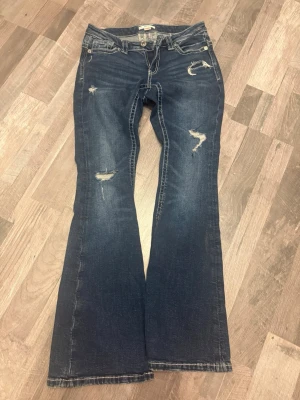 Lågmidjade jeans från Gina  - Snygga mörkblå jeans med vita kontrastsömmar och broderade detaljer på bakfickorna. Jeansen har slitningar framtill för en trendig look och klassisk femficksmodell. Perfekta för dig som gillar en avslappnad men ändå stilren stil.
