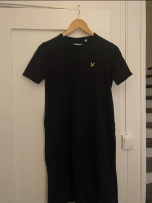 Svart t-shirtklänning från Lyle & Scott - Svart t-shirtklänning från Lyle & Scott i 100% ekologisk bomull. Klänningen har korta ärmar, rund halsringning och en liten gul logga på bröstet. Enkel och stilren modell som passar perfekt till sneakers eller sandaler.