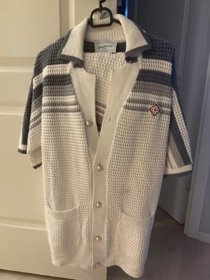 Casablanca sommarkläder set shorts och tröja stickad  - Kortärmad stickad kofta från Cashlance i vitt och grått med breda ränder på ärmarna och axlarna. Koftan har pärlemorknappar, två fickor fram och en liten broderad patch på bröstet. Snygg krage och grovstickad struktur ger en retrovibe.