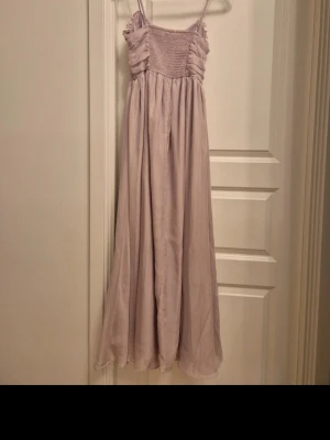 Ljuslila långklänning med smala band - Säljer en romantisk ljuslila långklänning med smala axelband och rynkad överdel. Klänningen har en luftig, svepande kjol och omlottdetaljer vid bysten. Perfekt för sommarens alla tillfällen! Använd 1 gång 