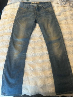 Wrangler ljusblå raka jeans - Säljer ett par ljusblå jeans från Wrangler med klassisk rak passform och snyggt slitna detaljer på framsidan. Jeansen har fem fickor, gylf med dragkedja och Wrangler-lapp på bakfickan. Tillverkade i bomull för en skön och avslappnad look.