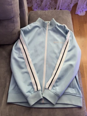 Ljusblå track jacket Palm Angels - Snygg ljusblå track jacket från Palm Angels med vita och svarta ränder längs ärmarna. Dragkedja framtill, hög krage och ribbade muddar. Tillverkad i ett glansigt syntetmaterial som ger en sportig vibe. Perfekt för dig som gillar streetwear och vill sticka ut.