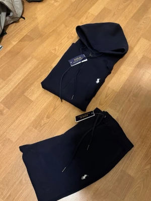 Mörkblått set från Polo Ralph Lauren storlek M inte använd - Snyggt mörkblått set från Polo Ralph Lauren med hoodie och matchande byxor. Båda delarna har den klassiska vita loggan broderad framtill. Setet är gjort i mjukt bomullsmaterial och har dragsko i både huvan och midjan för bästa passform.