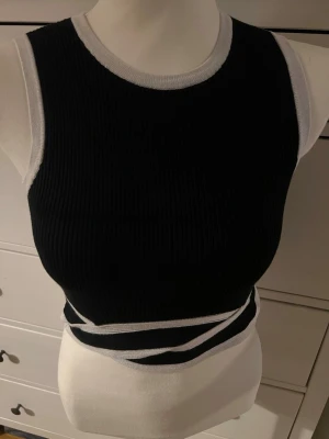 Svart och vit ribbad linnetopp - Snygg svart ribbad linnetopp med vita kontrastkanter och unika omlottdetaljer i midjan. Toppen är ärmlös och har en tight passform som framhäver figuren. Perfekt för dig som vill ha en trendig och stilren look.