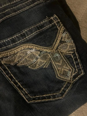 Mörkblå bootcut jeans med paljettdetalj - Säljer ett par mörkblå bootcut jeans med kontrastsömmar och unik broderad bakficka dekorerad med paljetter. Storlek 158