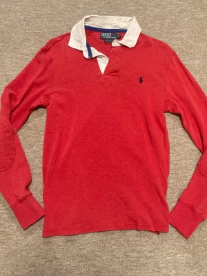Röd långärmad tröja Polo Ralph Lauren - Snygg röd långärmad tröja från Polo Ralph Lauren med vit krage och blå logga broderad på bröstet. Tröjan har förstärkta armbågar och ribbade muddar. Perfekt för en avslappnad men ändå stilren look.