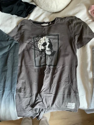 Grå t-shirt med dödskalle från KappAhl - Snygg grå t-shirt från KappAhl med tryck av en dödskalle och fåglar på bröstet. T-shirten har rund hals och korta ärmar. Materialet är mjuk bomull som känns skönt mot huden. Perfekt för dig som gillar lite edgy stil.