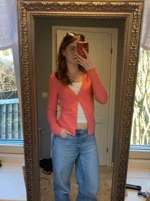 Korallrosa kofta - Korallrosa cardigan med v-ringning och långa ärmar. Tröjan har en enkel knappstängning framtill och är gjord i viskos och elastan. Enkel och stilren design som funkar till mycket. Tröjan har små deformiteter i sidsömmarna, och ett pytte hål i tygen precis vid lappen i nacken, annars inget fel.