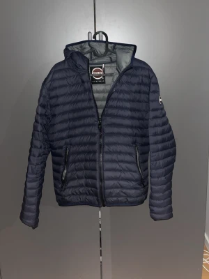 Mörkblå dunjacka från Colmar - Snygg mörkblå dunjacka från Colmar med huva och dragkedja framtill. Jackan har två fickor med dragkedja och quiltad design. Perfekt för kalla vinterdagar och har en stilren look med diskret logga på ärmen.