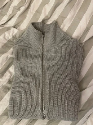 East west grå ziptröja med hög krage - Säljer en stilren grå stickad tröja med dragkedja framtill och hög krage. Tröjan har ribbstickad struktur och långa ärmar. Perfekt för lager på lager och enkel att matcha till olika outfits.