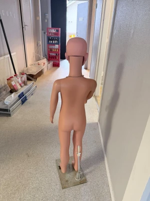 Barnmannequin i plast på metallplatta - Barnmannequin i hudfärgad plast med avtagbart huvud och armar. Står på en rektangulär metallplatta och har ett pärlhalsband runt halsen. Perfekt för att visa barnkläder eller accessoarer i butik eller hemma.