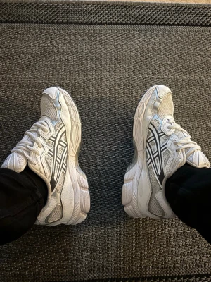 Vita Asics sneakers med mesh och detaljer - Säljer ett par vita Asics sneakers med klassiska svarta och grå detaljer. Skorna har en chunky sula och är tillverkade i mesh och syntetmaterial för en sportig look. Perfekta för dig som gillar retrostil och bekväma skor.