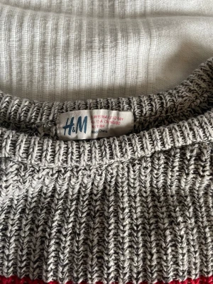 Randig stickad tröja från H&M - Stickad tröja från H&M med breda blockränder i grått, rött och mörkblått. Tröjan har rund halsringning och långa ärmar. Perfekt för kyliga dagar och enkel att matcha med jeans eller kjol.